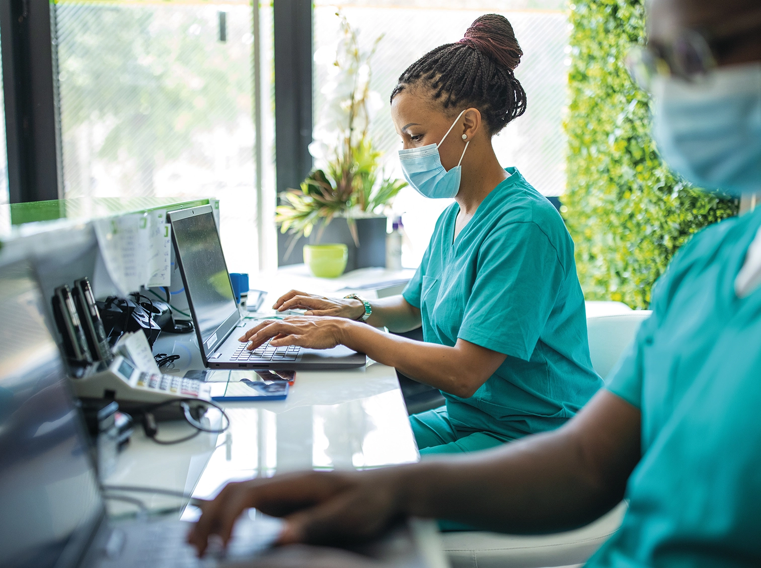 Healthcare workers in masks using laptops at a clinic desk.