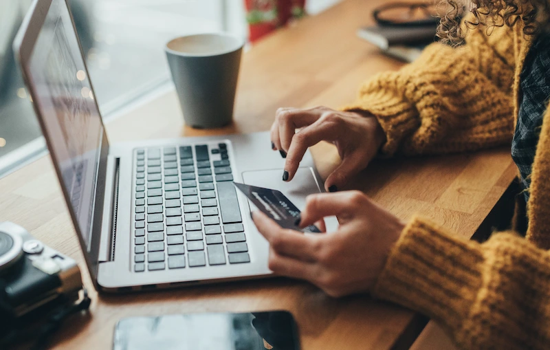 Person wearing a yellow sweater using a laptop and holding a credit card while shopping online.