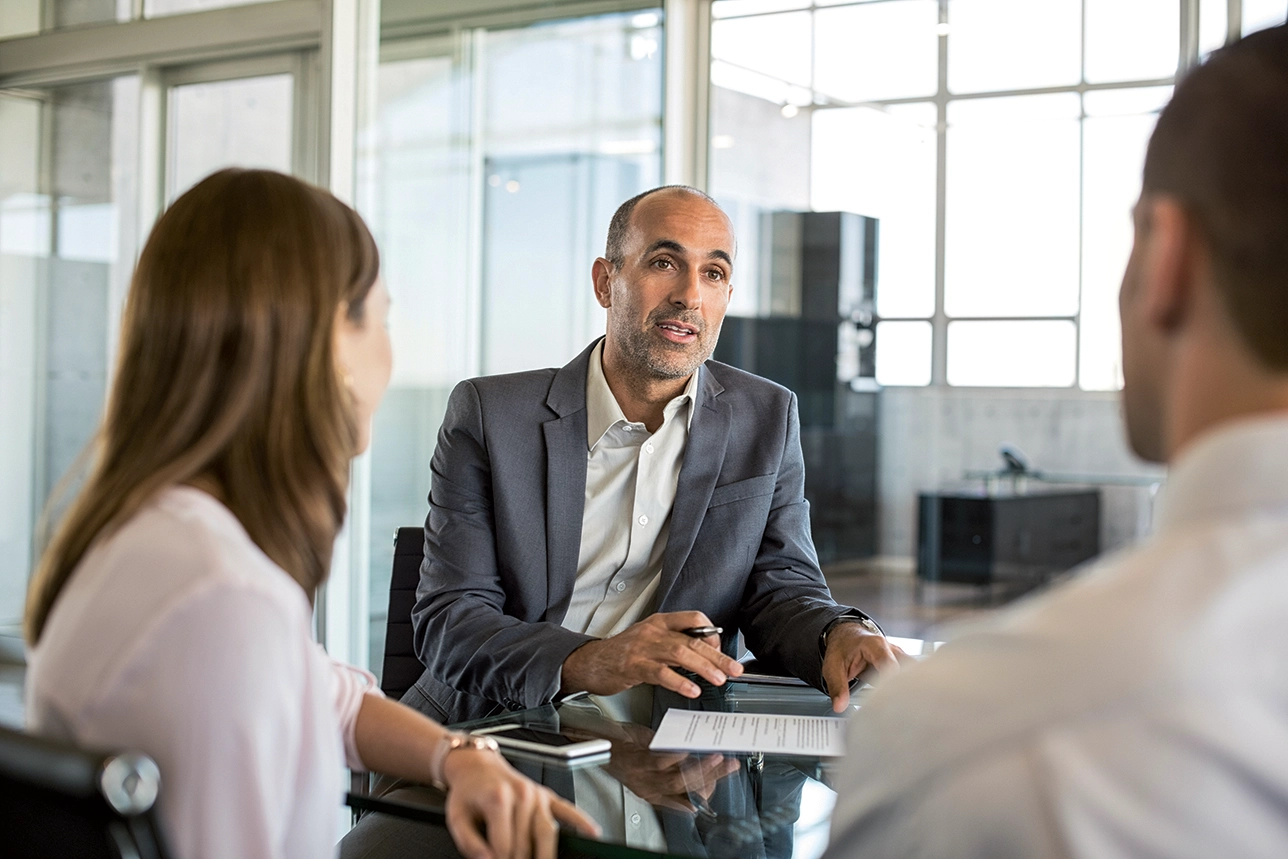 Business advisor speaking with two people at a glass table, pen in hand over a document in a bright office.