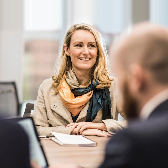 Smiling woman wearing a scarf and coat sitting at a table during a professional meeting.